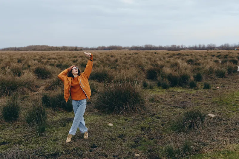 Woman outdoors field landscape walking, orange jacket and light denim, arms raised in joyful pose, grass and shrubs, cloudy sky, wide countryside view, lifestyle moment