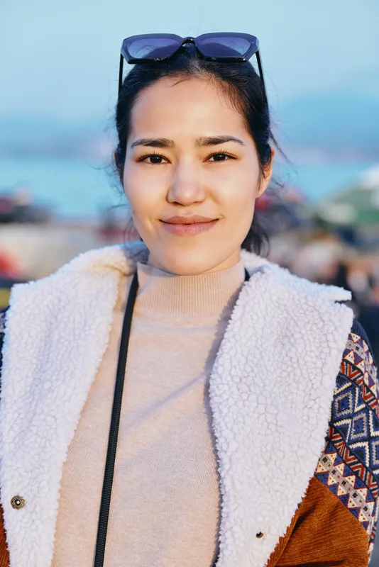 Woman in cozy sweater and jacket with sunglasses on head, outdoor fashion scene by seaside, natural light, relaxed pose, soft textures, casual lifestyle vibe, candid style suitable for fashion