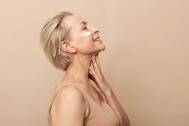 Mature woman with short blond hair applies face cream on cheek and smiles gently. Skincare concept focused on natural beauty and healthy skin care routine in soft beige studio setting.