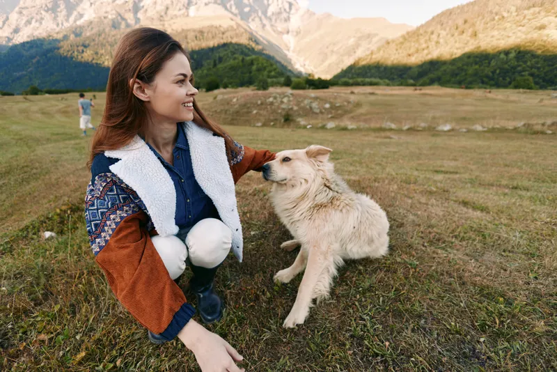 Woman crouching in a mountain field smiling while petting a dog, enjoying nature and companionship in a scenic outdoor landscape, casual jacket, happy moment with her pet