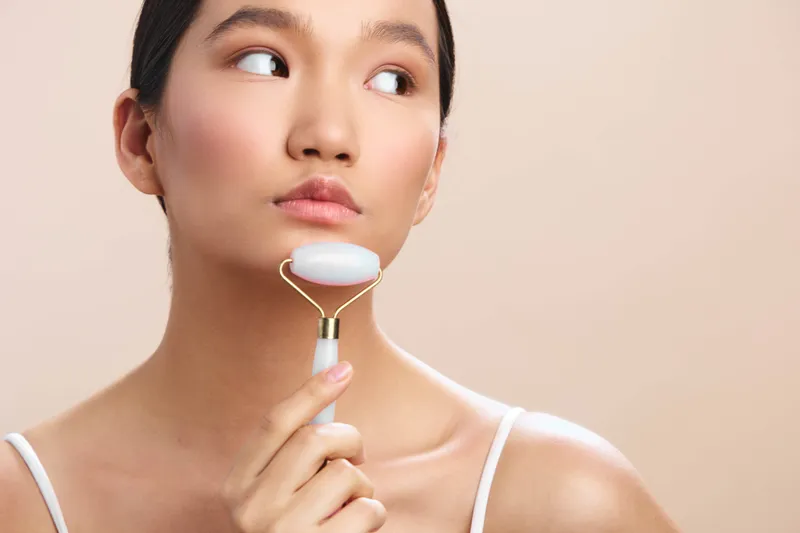 asian woman uses facial roller in a skincare routine, close up shot with soft lighting, calm expression and clean aesthetic, serene spa vibe, minimal background, healthy glow and selfcare mood.
