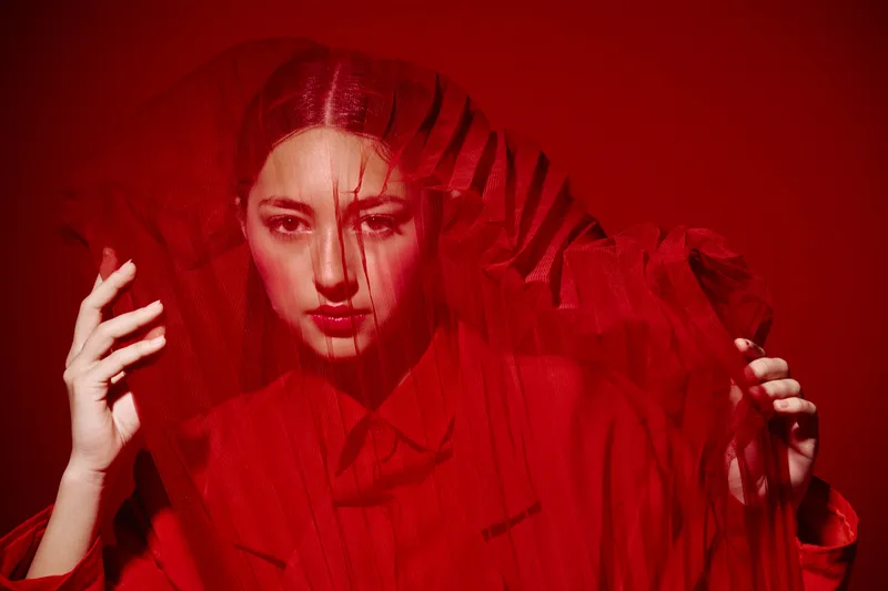 Portrait of a creative woman in bright red fashion with a sheer red veil hat. Studio lighting with strong red background enhances artistic and avant-garde beauty concept.