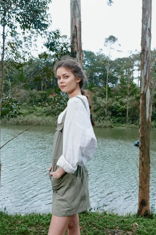 Young woman in overalls by a serene lake, showcasing natural beauty, casual style, and a contemplative mood amidst lush greenery