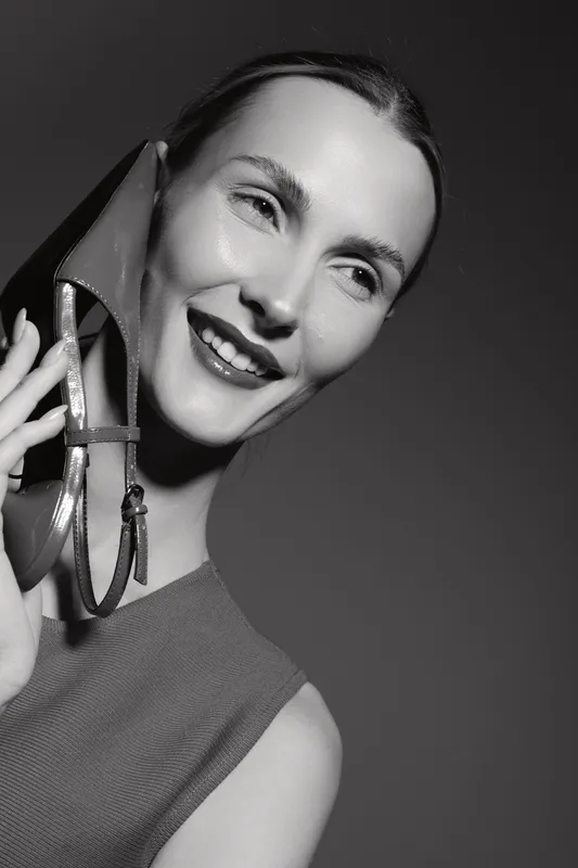 Smiling Caucasian woman with light skin holds high heel shoe near face in monochrome studio portrait. Concept of fashion, beauty, and elegance.