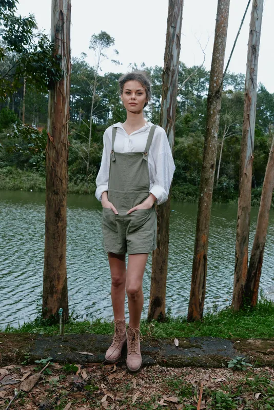 Young woman in green overalls standing by a serene lake, showcasing a casual outdoor style, peaceful emotions, and a natural, earthy background