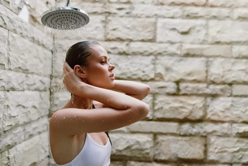 Woman taking outdoor shower under rain showerhead with wet hair, stone wall background, natural light, casual white top, calm expression, refreshing moment.