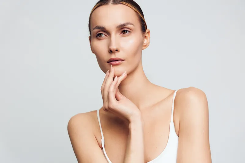 Thoughtful woman with clear skin posing with hand on chin, neutral expression, natural makeup, wearing white top, studio lighting, clean background, minimalistic style