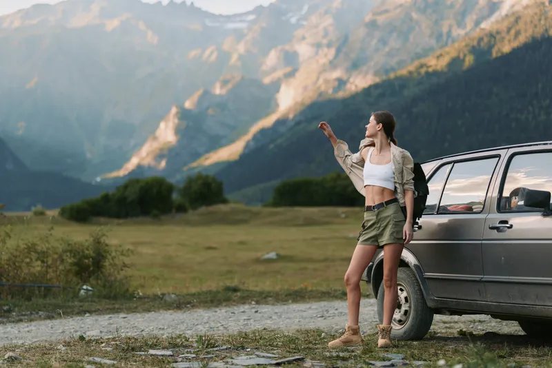 A young woman in a white crop top and green skirt stands next to a car on a mountain road, looking into the distance with a relaxed and confident pose.