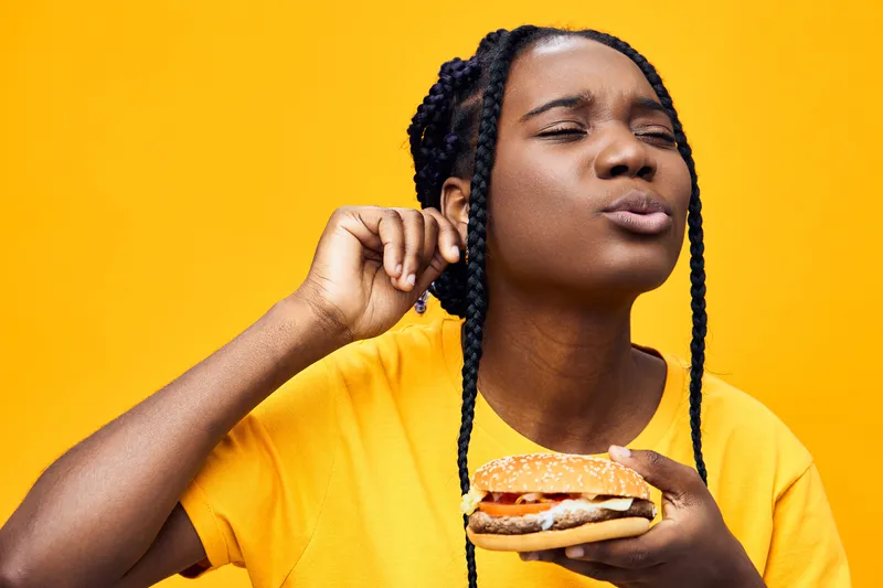 Young woman enjoying a delicious hamburger with a joyful expression against a vibrant yellow background, highlighting the concept of indulgence in fast food