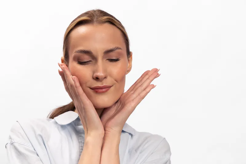 Young woman with clear skin and closed eyes gently touches her face with both hands. Concept of skincare, natural beauty, self-care, and wellness on white background.