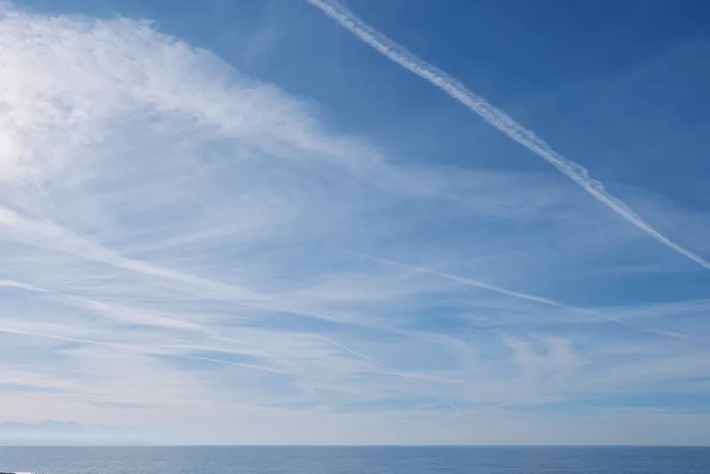 Clear blue sky over a calm ocean, intersecting wispy clouds and contrails, expansive horizon, serene seascape perfect for travel, nature, and outdoor mood in bright daylight.
