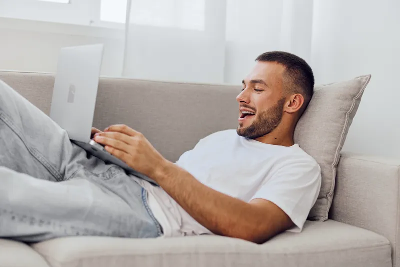 Smiling man working on a laptop, relaxed at home. Comfortable setting with a cozy sofa and natural light. Ideal for modern lifestyle themes in 2025.