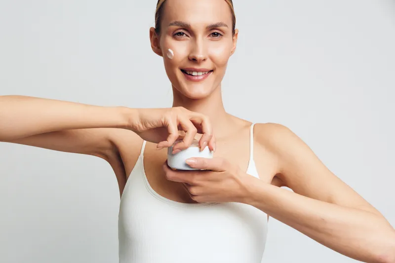Smiling woman applying white face cream on her cheek. Close-up on a light background. Skincare, self-care, wellness concept. Soft studio lighting creates a warm, calm mood.