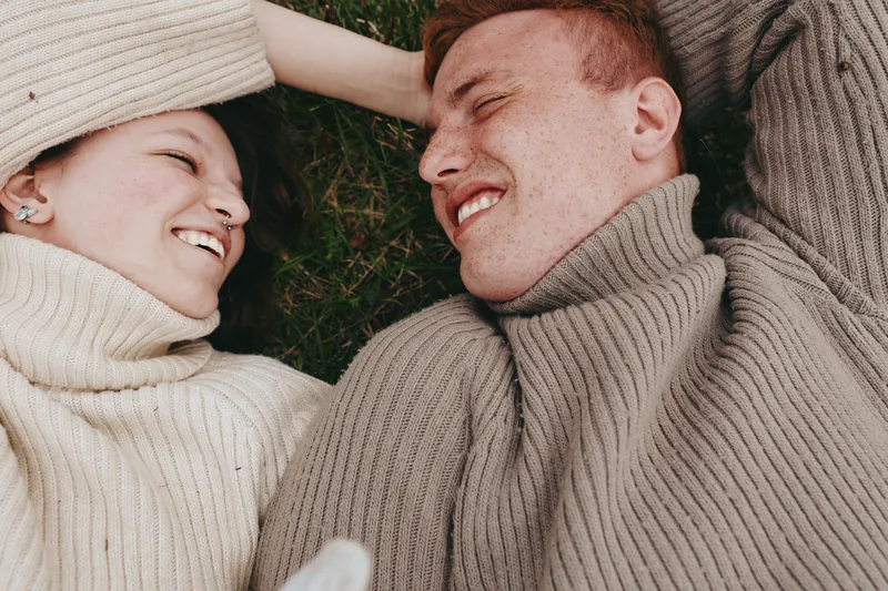 Smiling couple lying on grass in matching beige sweaters, relaxed embrace, warm sunlight. Concept of lifestyle, comfort, and romance. Close-up on soft textures and natural setting.