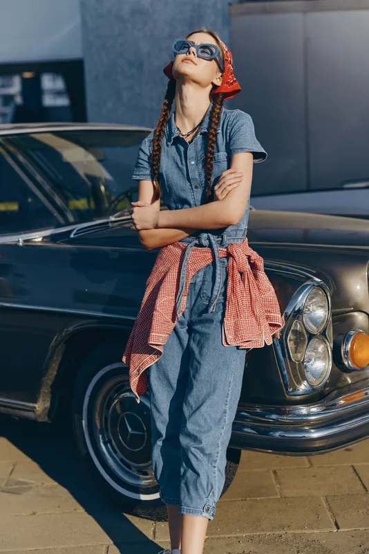 Lifestyle woman in modern boho-western denim street style with sunglasses and braided hair, film color aesthetic, relaxed pose near vintage car during daytime.
