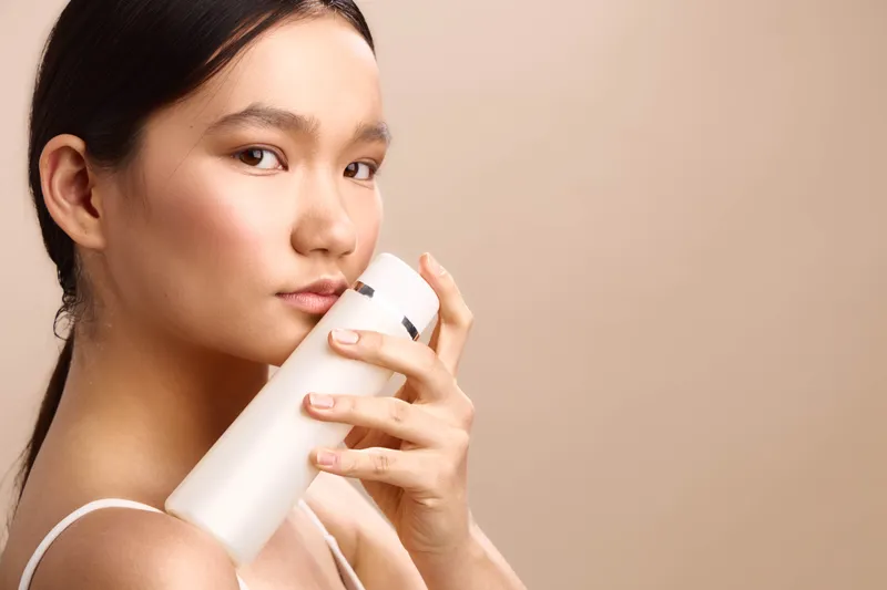 Asian woman in a soft studio holds a sleek skincare bottle, showcasing a calm, focused expression. The intimate closeup highlights luminous skin and a serene beauty moment.