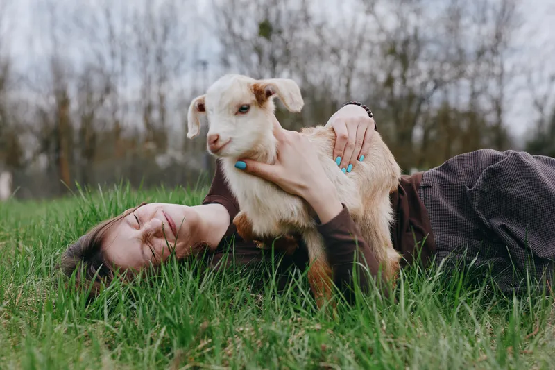 Goat, kid, woman outdoors, lifestyle moment on grass with gentle cuddle, joyful expression, sunlit calm, relaxed posture, natural colors, lush background, rural setting, affectionate interaction with