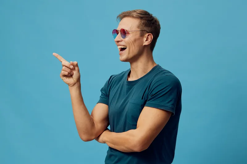 Smiling man wearing sunglasses, casual blue t-shirt, pointing to the side and looking happy, isolated on solid bright blue background. Casual lifestyle concept, joyful expression, fun mood, positive a