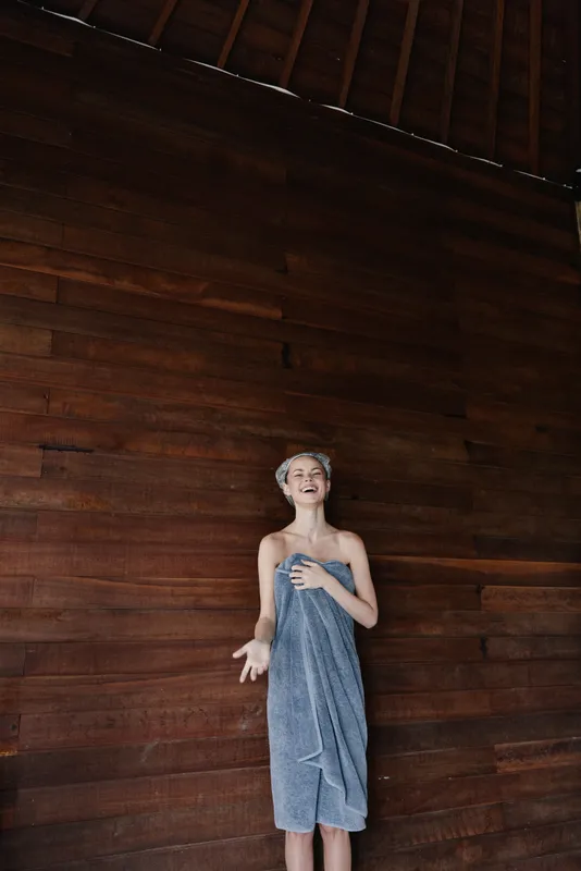 smiling woman wrapped in grey towel with hair rollers standing against dark wood wall, natural light, relaxed mood, indoor setting, skincare routine moment