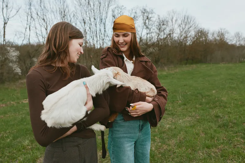 Two women outdoors with a kid goat, sharing a gentle moment in a grassy field. Casual fashion, rustic vibe, and animal interaction convey warmth, nature, and everyday countryside life.