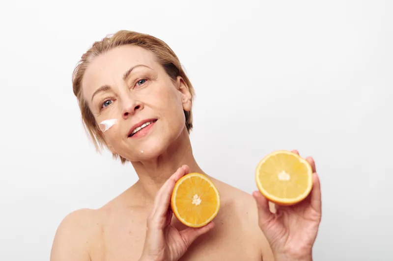 Mature woman with facial cream on cheek holds two orange halves smiling on white background. Concept of natural skincare, healthy lifestyle, vitamin c, and beauty care for aging skin.