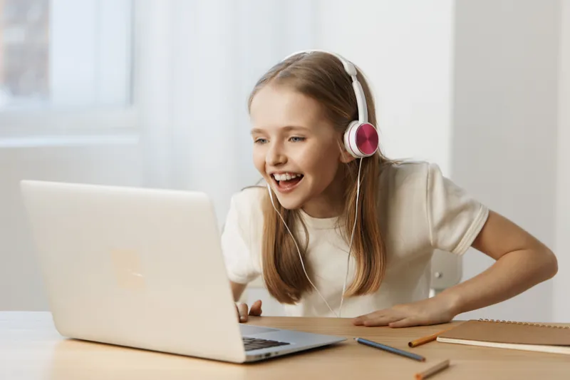 Happy girl in white t shirt with headphones, excitedly using a laptop for online learning or gaming in a bright room with soft lighting