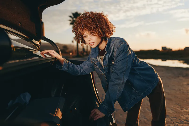 Woman lifestyle outdoors by sunset, curly hair girl leaning on car door, denim jacket, warm golden light, relaxed confident pose near water, soft breeze, cinematic background
