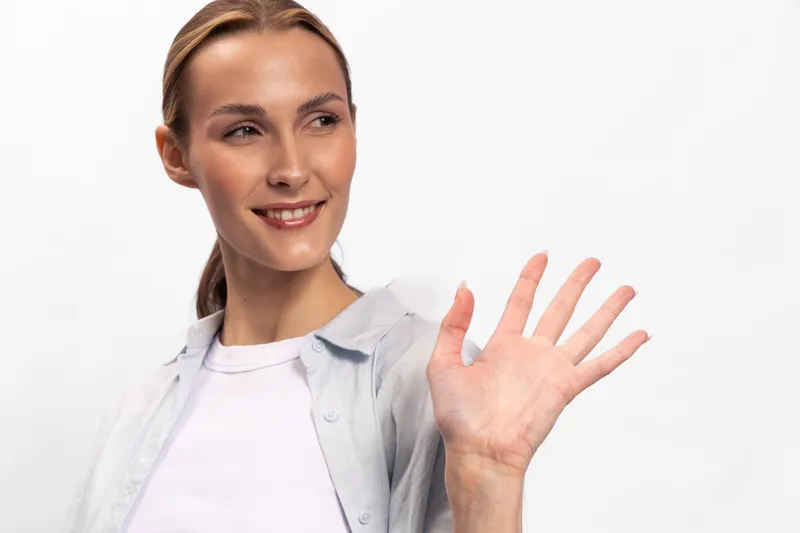 Smiling young woman with light skin waves hand in greeting, wearing casual white shirt and light jacket. Bright studio background emphasizes friendly and positive mood concept.