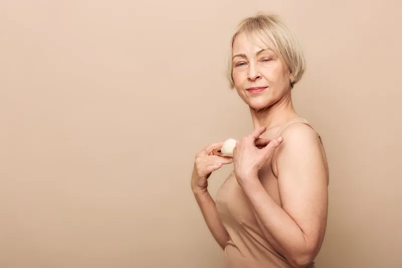 Confident mature woman applies natural deodorant on shoulder with relaxed expression, wearing beige tank top. Concept of personal care, hygiene, and natural skincare for older adults.