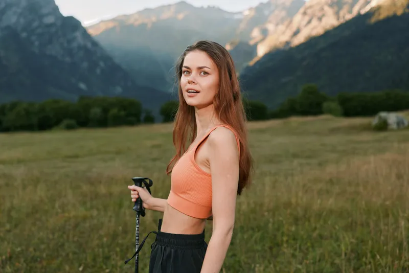 A young woman stands in a wide grassy meadow with hiking poles, wearing a coral crop top and dark pants. She faces the camera with a calm smile as distant mountains rise.