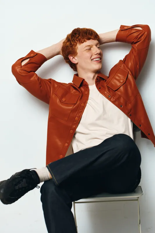 Young man with red hair smiling and sitting relaxed on chair wearing white shirt and brown jacket with black shoes casual style against white background.