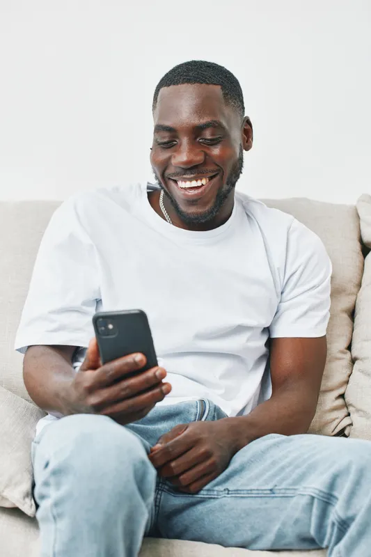 Happy young African man using smartphone on a sofa, showcasing a relaxed lifestyle and modern technology in a cozy interior setting