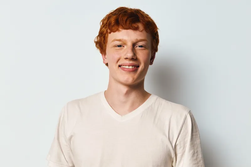 Young man with red hair and freckles smiling and posing in casual white t shirt on light background. Portrait of cheerful male adult with clear skin and friendly expression.