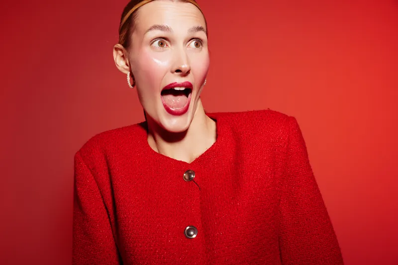 Surprised woman in red jacket with open mouth looks aside on red background. Concept of emotion, excitement, and fashion portrait with bright studio lighting.