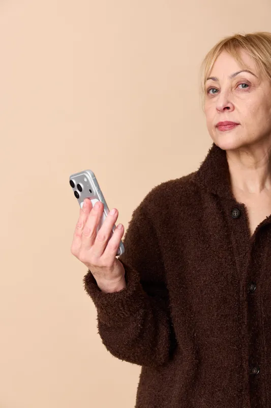 Senior woman in dark sweater holds a smartphone, worried face, indoor beige background, portrait concept for anxiety, loneliness, and technology communication