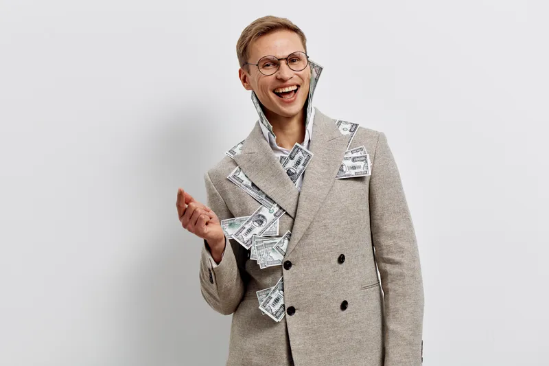 laughing young caucasian man wearing beige coat with money bills in pockets and joyful expression on white background digital nomad concept