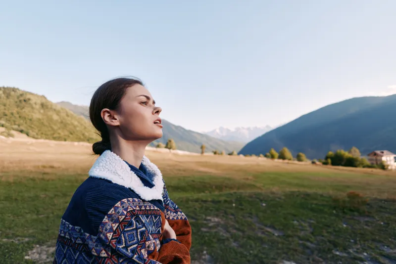 Woman portrait in nature on a mountain meadow wearing a patterned sweater, enjoying fresh air outdoors and travel vibe with peaceful expression and scenic valley landscape.