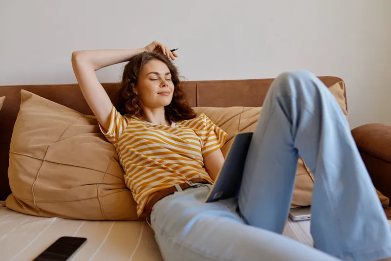 Young woman relaxes on a sofa at home with a laptop, eyes closed, content smile, casual striped tee and light jeans, cozy indoor moment