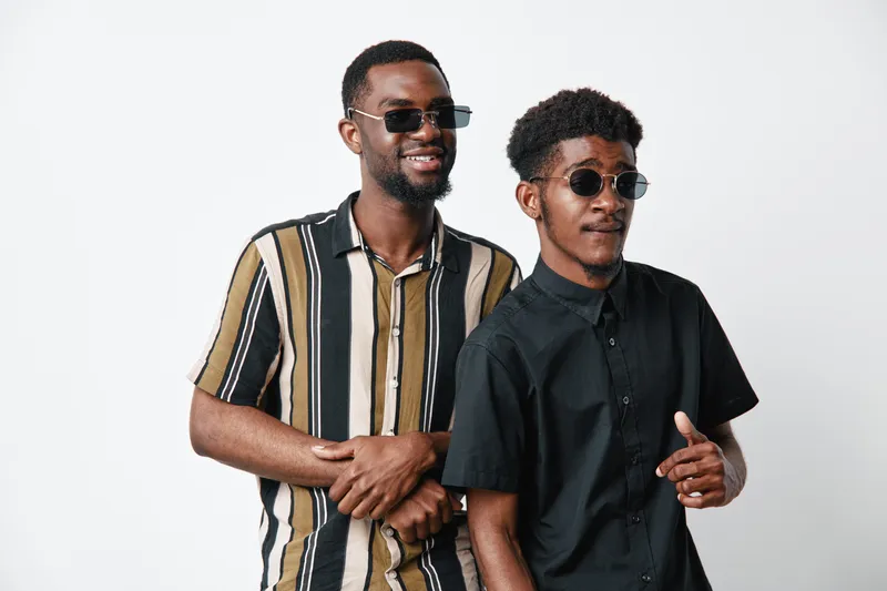 Two African men with dark skin wearing sunglasses and casual shirts standing together on white background smiling and posing for portrait photography with confident expressions.