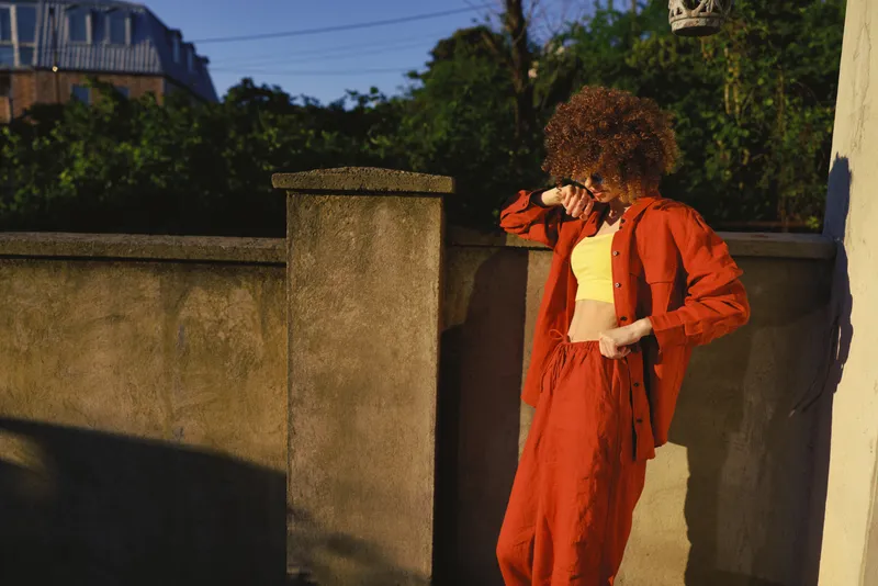 A stylish woman in a bold red outfit stands by a sunlit wall, adjusting her sleeve. A candid outdoor fashion moment highlighting lifestyle, confidence, casual street style, natural light, fashion