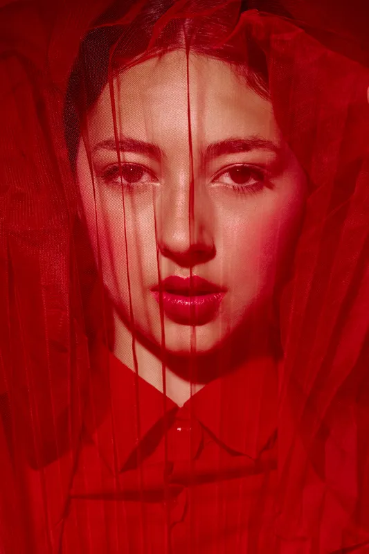 Close-up portrait of a young woman in red makeup and clothing, partially veiled with red fabric. Creative fashion beauty shot emphasizing bold color and dramatic mood lighting.