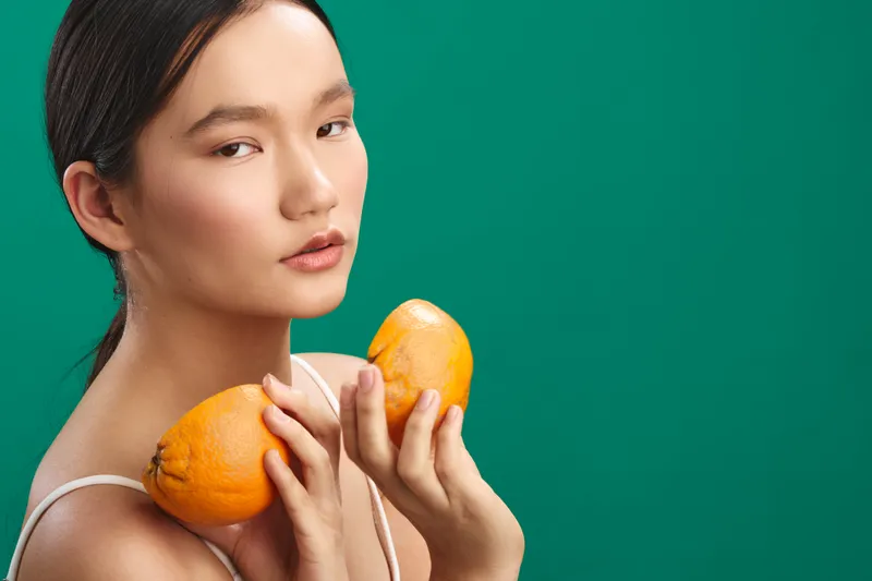 Asian woman holds bright oranges against a teal background, presenting a fresh and vibrant lifestyle moment. Her composed expression and soft lighting convey wellness and vitality.