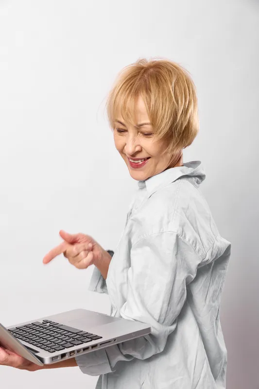 Senior woman using laptop typing for work and smile in an online call, white studio background, cheerful laughing expression, modern technology and productivity