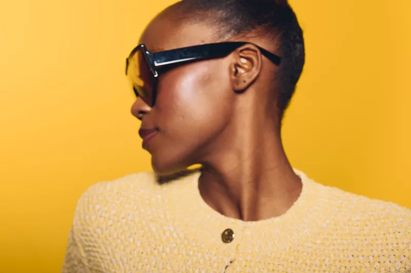 Profile of a dark skinned African woman wearing sunglasses and a yellow textured sweater against a bright yellow background. Concept of fashion, style, and confident beauty.