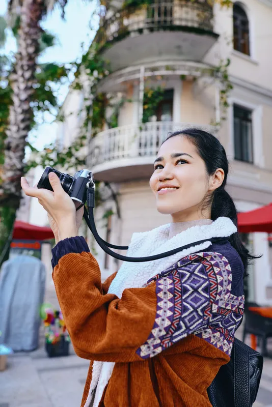 Camera in hand, woman outdoor street photography joy, smiling while exploring a sunny plaza with colorful awnings and leafy backdrop, capturing candid urban moments and vibes