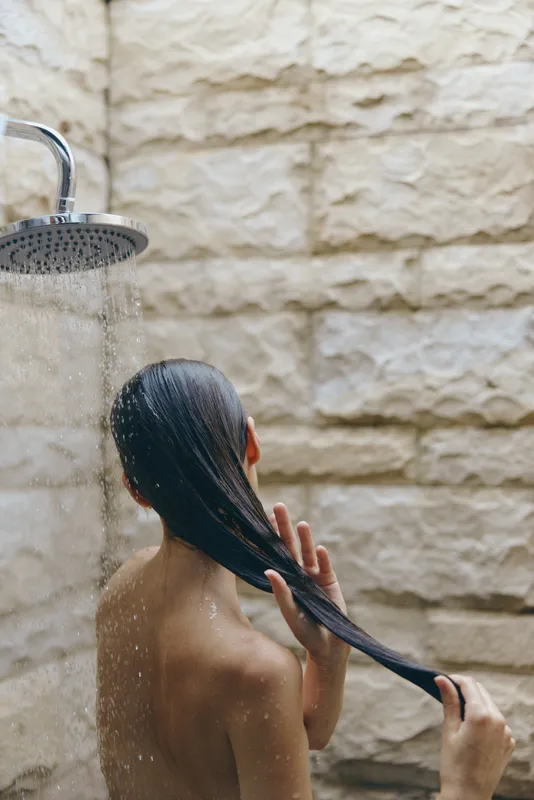 woman washing long black hair under outdoor shower with stone wall background, natural skincare routine in warm sunlight, peaceful atmosphere