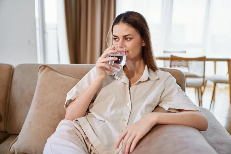 Woman on sofa drinking water from glass at home, relaxed and thoughtful on couch in livingroom, casual clothing and natural light, weekend rest and hydration routine.