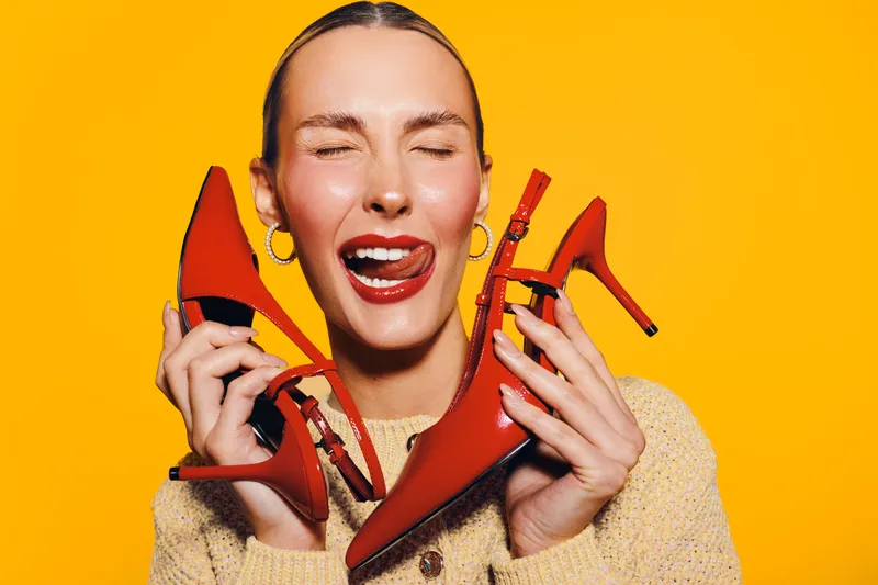 Happy woman with closed eyes holds red high heels near face, smiling with tongue out. Bright yellow background creates vibrant mood. Concept of fashion, shoes, and joyful emotions.