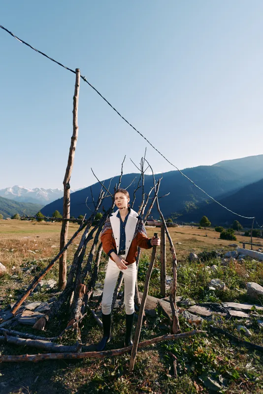 Person wearing shearling jacket and boots stands beside rustic fence in mountain field, countryside landscape with clear sky and distant peaks, outdoor fashion portrait.