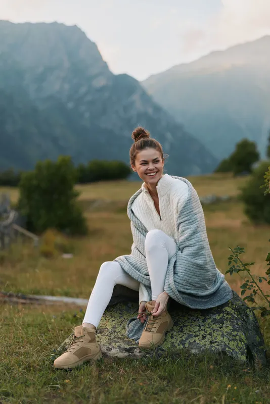 Young woman sits on a mossy rock in a mountain meadow, adjusting her hiking boot and smiling softly as warm light highlights her relaxed, hopeful mood.